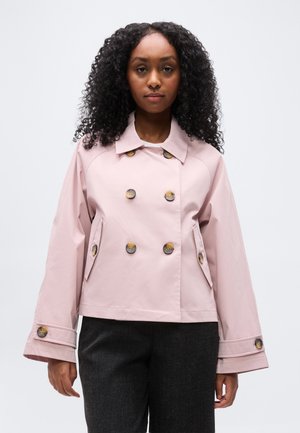 Žena s kučeravými vlasmi v svetloružovom dvojradovom saku s veľkými gombíkmi a tmavými nohavicami na jednoduchom pozadí.