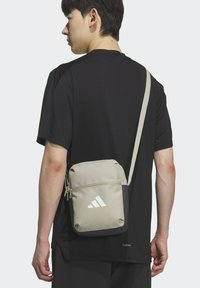 Bežová crossbody taška so zipsovým uzáverom, na ktorej je jemné logo Adidas. Taška je vyrobená z kvalitného materiálu s hladkou textúrou.
