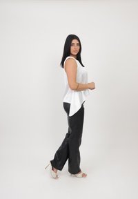 Witte mouwloze top met een gebogen zoom, gecombineerd met strakke zwarte leren broek en gepatternde haksandalen. Effen witte achtergrond.