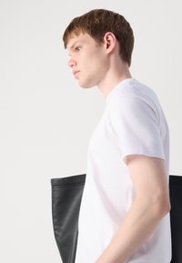 White short-sleeve t-shirt, smooth cotton material. Model holds a large black leather tote bag with a soft texture and rounded corners.