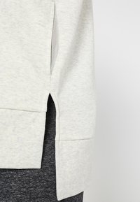 Ourlet de sweat-shirt gris clair avec une fente latérale révélant la doublure en mesh noir de la poche et un pantalon gris foncé chiné en dessous.