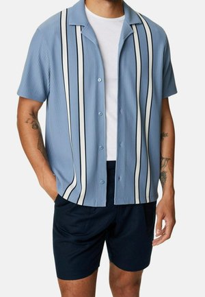 Chemise bleu clair à manches courtes avec des rayures verticales blanches et noires, en tissu texturé, associée à un short navy et un T-shirt blanc.