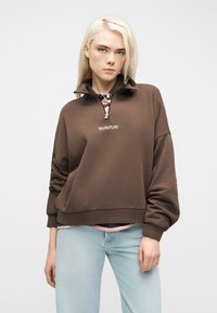 Ξανθιά γυναίκα με καφέ φούτερ με φερμουάρ στη μέση και το κείμενο "NAPAPIJRI" καθώς και ανοιχτόχρωμα μπλε τζιν, στέκεται μπροστά σε λευκό απλό φόντο.