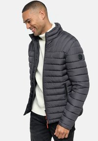 Chaqueta de plumas gris con acolchado vertical, cuello alto y bolsillos con cremallera. Cuenta con un parche de logo en el brazo izquierdo y detalles en cremallera naranja.