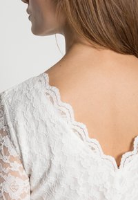 Haut en dentelle blanche avec encolure festonnée et motifs floraux, révélant une texture dorsale lisse avec des détails complexes.