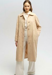 Palton lung bej cu guler, decorat cu mici embellishments, având o croială dreaptă și nasturi frontali, purtat peste o bluză albă și pantaloni.