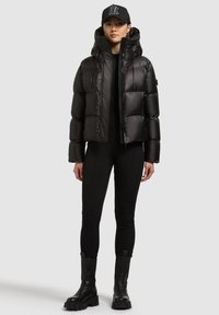 Musta untuvatakki, jossa on huppu, vetoketjusuljenta, joustavat hihat ja kiiltävä pinta. Päällä mustat istuvat vaatteet ja paksupohjaiset saappaat.