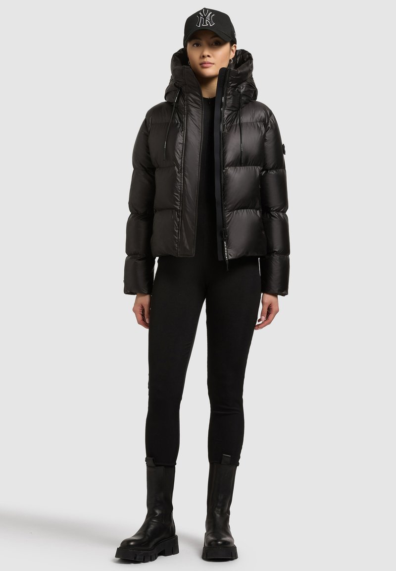 Musta untuvatakki, jossa on huppu, vetoketjusuljenta, joustavat hihat ja kiiltävä pinta. Päällä mustat istuvat vaatteet ja paksupohjaiset saappaat.