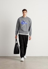 Mann iført grå sweatshirt med blå grafikk, svarte bukser, hvite sneakers, bærende svart totebag, som går mot en enkel hvit vegg i bakgrunnen.