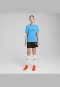 Gaiši zils Puma krekls ar teksturētu dizainu, melni šorti, balti zeķes un spilgti oranži futbola apavi. Uz krekla un zeķēm ir logo.