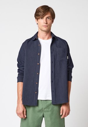 Jeune homme portant une chemise à rayures bleu marine à boutons par-dessus un t-shirt blanc et un pantalon vert, debout devant un fond uni.