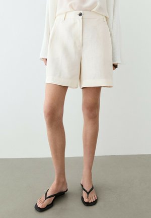 Person trägt cremefarbene maßgeschneiderte Shorts, weißes Oberteil und schwarze Flip-Flop-Sandalen, steht auf einem grauen Boden vor einer weißen Wand.