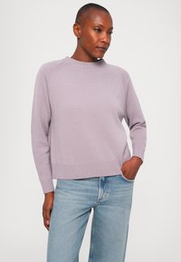 Woman with short hair wearing a light purple knit sweater and light blue jeans with one hand in pocket, standing against a plain background.