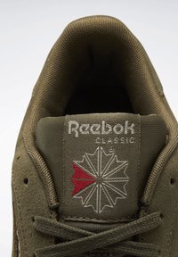 Sneaker Reebok Classic verde oliva con linguetta che mostra il logo ricamato, accompagnato da un design geometrico rosso e bianco e lacci coordinati.