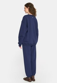 Kobieta z kręconymi włosami, ubrana w granatową, luźną bluzę oraz pasujące dresy, stoi zwrócona w lewo na jednolitym tle.