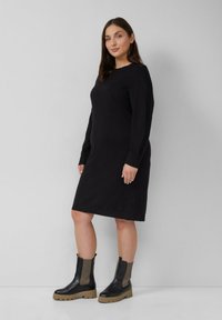 Frau, die ein schwarzes langärmeliges knielanges Kleid trägt, kombiniert mit schwarzen und beige Klobigen Stiefeln bis zu den Knöcheln, steht vor einer grauen Wand.