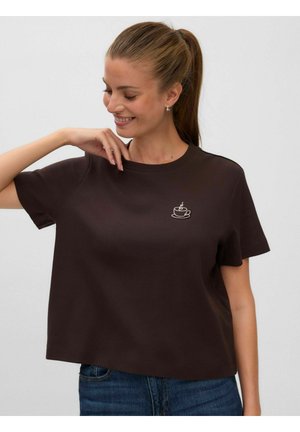 Femme aux cheveux bruns en queue de cheval portant un t-shirt marron foncé à manches courtes avec un petit motif blanc de tasse à café, souriante et regardant vers le bas.