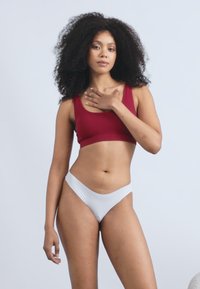 Červený rebrovaný crop top skombinovaný s bielymi nízkymi plavkami, ktoré majú hladké textúry a priliehavý strih. Model pózuje na neutrálnej pozadí.