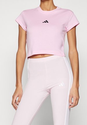 Kobieta w jasnoróżowej krótkiej koszulce i dopasowanych leginsach w tym samym kolorze, oba z małymi logo Adidas i białymi paskami po bokach leginsów.