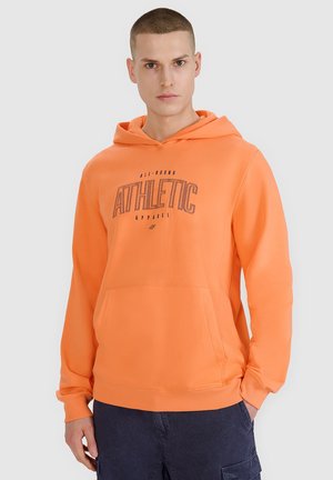 Młody mężczyzna z krótkimi włosami, ubrany w pomarańczową bluzę z kapturem z napisem "ATHLETIC" oraz ciemnoniebieskie spodnie typu cargo, stoi na jasnym tle.