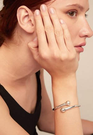 Bracelet en argent présentant un design lisse et arrondi avec deux accents sphériques. Porté au poignet d'une personne avec un arrière-plan neutre.