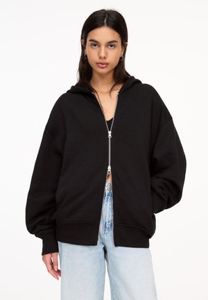 Sort zip-up hoodie med en stor, afslappet pasform, forkromet med hætte og ribbede manchetter. Sammen med lyseblå denim jeans.