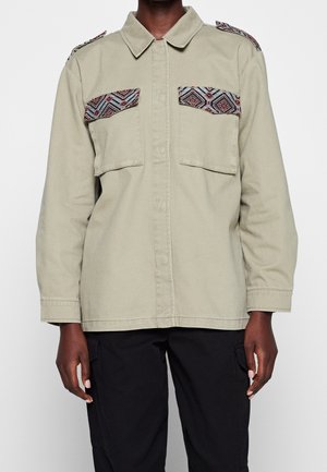 Personne portant une veste beige à manches longues avec des poches poitrine à motifs et des épaulettes, associée à un pantalon noir sur fond uni.