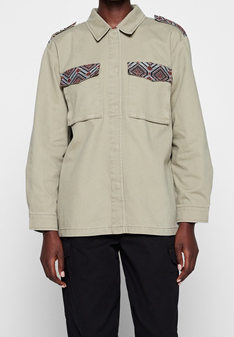 Personne portant une veste beige à manches longues avec des poches poitrine à motifs et des épaulettes, associée à un pantalon noir sur fond uni.