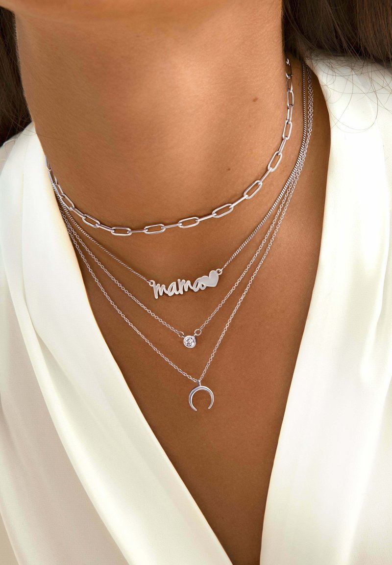 Collane a strati in argento con una catena a maglie, un pendente "mama", una gemma e una luna crescente, tutte poggiate sulla pelle.