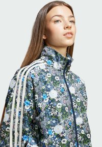 Blommönstrad zip-jacka med hög krage, blåa och gröna färger, samt vita accentränder på axlarna. Lättviktsmaterial.