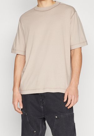 Uomo che indossa una maglietta beige a maniche corte e pantaloni cargo neri con tasche, in piedi davanti a uno sfondo chiaro e semplice.