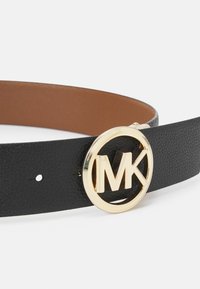 Svart läderbälte med en slät yta, prytt med en gyllene runt spänne med "MK"-logotyp. Insidan har en ljusbrun finish.