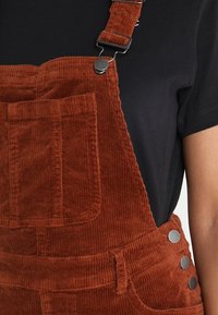 Salopette en velours côtelé couleur rouille avec bretelles réglables, détails en métal argenté, poche avant et tissu texturé.