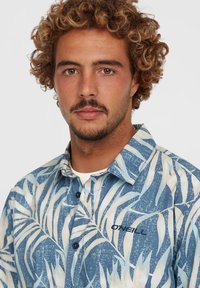 Camisa de manga corta con botones que presenta un patrón de hojas en azul y crema. El material es de tela ligera, con un logo de la marca en el pecho.