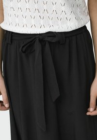 Pantalons larges noirs avec une taille plate et une ceinture en tissu, présentant une texture douce et un design fluide, associés à un haut en tricot blanc.