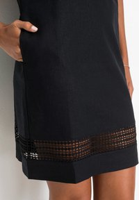 Schwarzes Kleid mit einem strukturierten Saum und geometrischen Ausschnitt-Details. Verfügt über seitliche Taschen für zusätzliche Funktionalität. Glatte Stoffoberfläche.