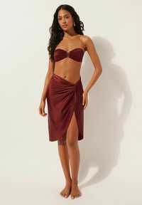 Bordeauxrotes trägerloses Bikinioberteil mit goldfarbenem Akzent, kombiniert mit einem passenden Wickelrock mit Drehknoten und seitlichem Schlitz. Glatte Textur.