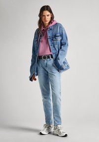 Chaqueta de mezclilla azul claro sobre un hoodie rosa, combinada con jeans azul claro y zapatillas grises. Presenta un cinturón negro y un diseño con capucha.