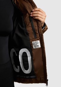 Braune, gesteppte Jacke, offen, zeigt ein schwarzes Innenfutter mit großen weißen Buchstaben und einem Etikett mit der Aufschrift "Eine warme Umarmung aus Stoff."