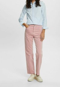 Femme portant une chemise à manches longues bleu clair, un pantalon droit rose, des baskets blanches, une main dans la poche et des bagues aux doigts.