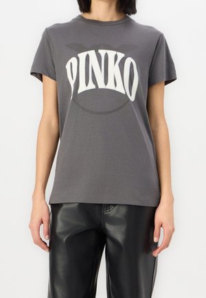 Persoon draagt een grijs T-shirt met korte mouwen met "PINKO" in grote witte letters en een zwarte leren broek, te zien van nek tot heupen.