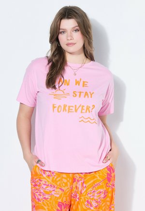 Kobieta o długich brązowych włosach nosi różowy T-shirt z napisem „Czy możemy zostać na zawsze?” oraz pomarańczowe spodnie w kwiaty, stoi przy białej ścianie.