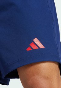 Marina atletiska shorts med en texturerad tyg och en röd Adidas-logotyp som accent vid fållen. Skräddarsydd passform med en mjuk inre foder.