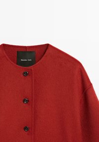 Cappotto rosso in lana con scollo arrotondato, caratterizzato da una pattina frontale con tre bottoni neri e una superficie morbida e strutturata. Etichetta visibile.