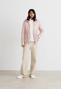 Chaqueta rosa claro con cremallera y capucha, con cintura elástica y bolsillos laterales, llevada sobre un suéter blanco y pantalones beige. Zapatillas blancas.