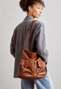 Brown leather bucket bag featuring two front pockets with buckle accents. The bag has a smooth texture and an adjustable strap.