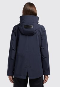 Giacca impermeabile blu navy con colletto alto, cappuccio regolabile e pannello posteriore texturizzato con logo. Cuciture visibili e chiusure a pressione.