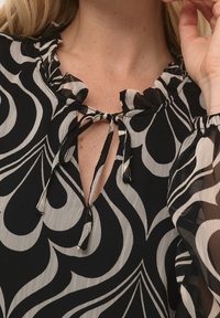 Blouse à motif noir et blanc avec un col froncé, un détail à nouer et de longues manches transparentes ornées de motifs tourbillonnants.