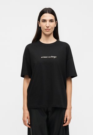 Kvinde iført en sort oversized T-shirt med teksten "armani exchange" på brystet, stående med arme afslappet mod en ensfarvet hvid baggrund.