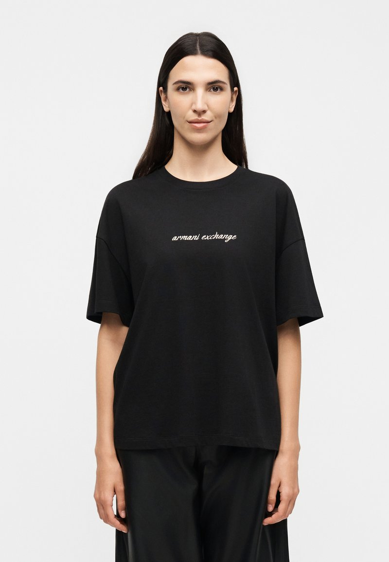 Γυναίκα που φοράει ένα μαύρο oversized T-shirt με το κείμενο «armani exchange» στο στήθος, στέκεται με χαλαρά χέρια μπροστά σε απλό λευκό φόντο.
